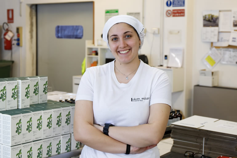 Samanta, staff member at Dott. Solari Cosmetics, wearing a hairnet and standing in the production area with finished product boxes.