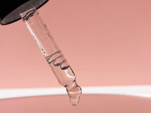 Clear cosmetic serum dripping from a glass dropper against a soft pink background.